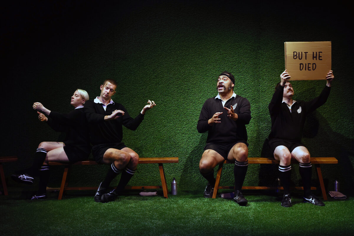 Four people in black athletic uniforms sit on a bench against a green wall; three mime actions, while one holds a cardboard sign reading BUT HE DIED. The scene is dramatic and theatrical.