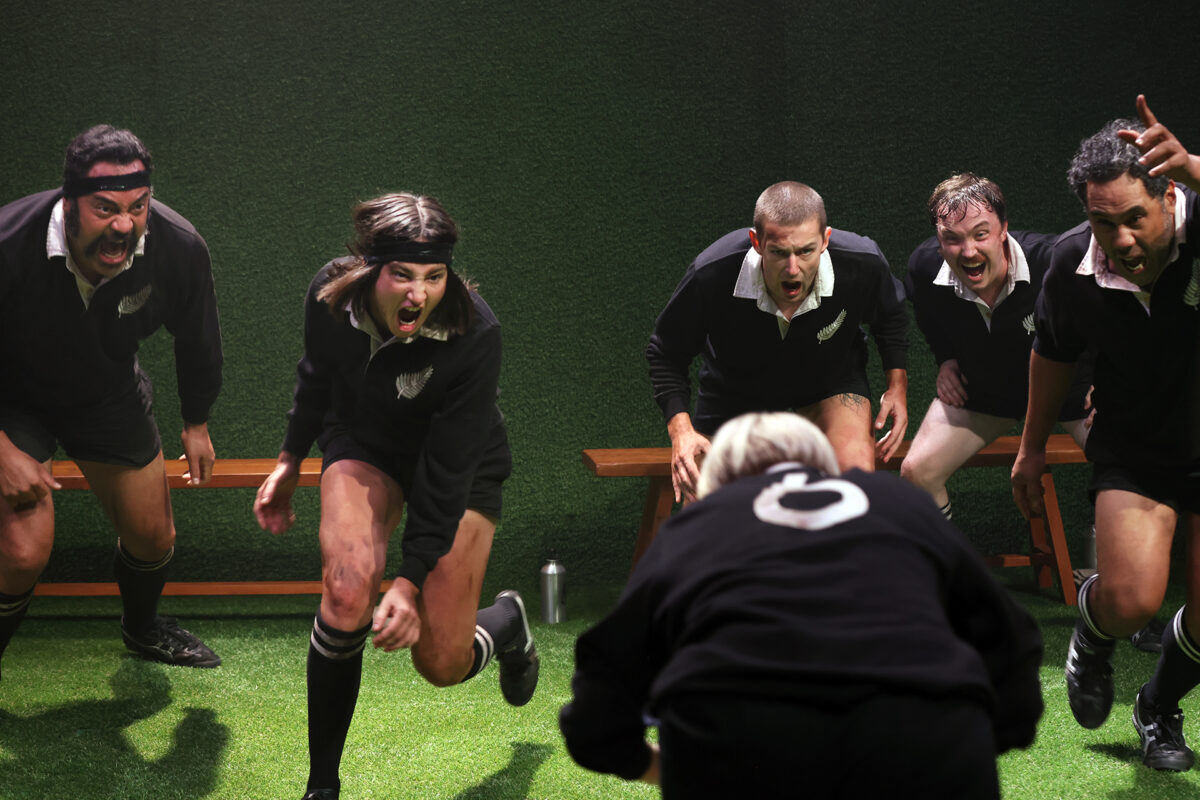 Five rugby players in black kits perform a fierce haka, shouting and gesticulating aggressively towards a person in a black shirt with the number 9, who stands with their back to the camera on green turf.