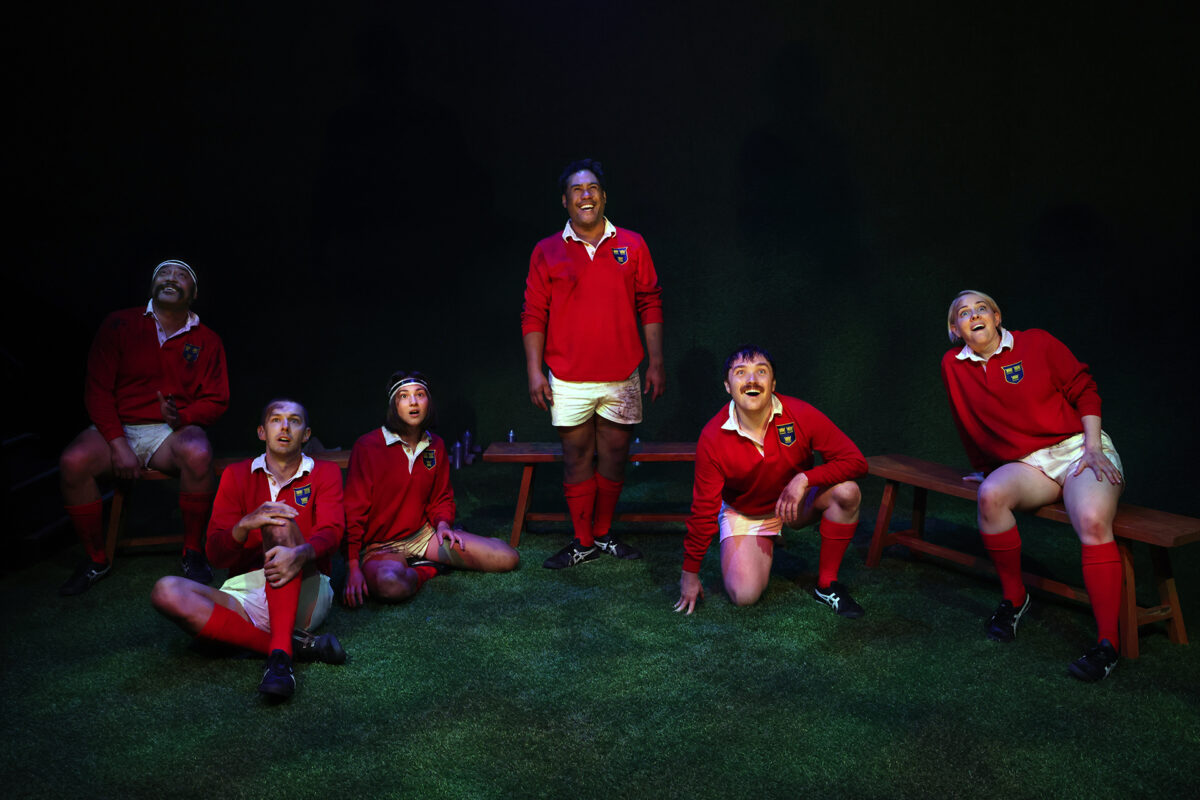 Six people wearing red rugby kits sit or kneel on artificial grass, looking up with expressions of surprise or awe. Wooden benches are on either side, and the background is dark and simple.