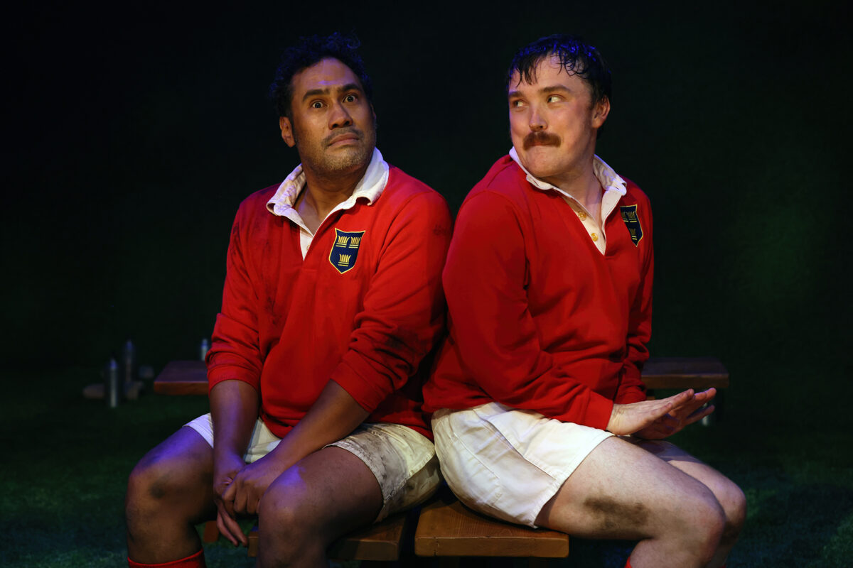 Two men in red rugby shirts and white shorts sit back-to-back on a bench, both with muddy legs. One looks up with a surprised expression, while the other looks sideways, smiling slightly.