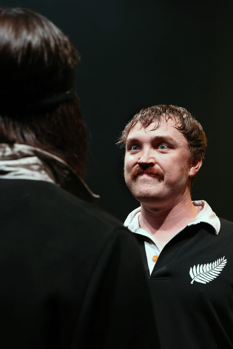 A man with a moustache and wide eyes stares intently at another person whose back is to the camera. The man is wearing a black shirt with a white fern emblem, standing under dramatic stage lighting.