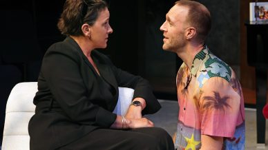 A woman in a black outfit sits on a white chair, holding hands with a man in a colourful, tropical shirt who kneels in front of her. They look at each other intently, appearing deep in conversation.