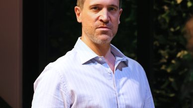 A man wearing a light blue striped shirt and khaki trousers stands indoors, holding a glass of white wine. He looks slightly to the side, with a neutral expression. There is a small framed photo on the table beside him.