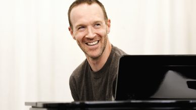 A smiling man with short hair and a light beard sits behind a black grand piano, looking slightly to the side. He is wearing a grey T-shirt and the background is plain and light-coloured.