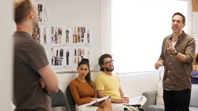 A group of four people are in a meeting room. One man stands and speaks whilst holding up two fingers. Two people sit, listening, and another man stands facing the group. Fashion sketches are displayed on a whiteboard behind them.