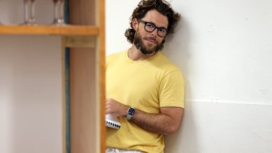 A man with glasses, a beard, and wavy hair leans against a white wall, wearing a yellow T-shirt and white trousers. He holds a notebook and looks at the camera, partially obscured by a nearby wooden shelf with glasses.