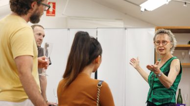 A woman with short grey hair and glasses gestures while speaking to three people in a casual indoor setting; one person holds a drink, and the group listens attentively.