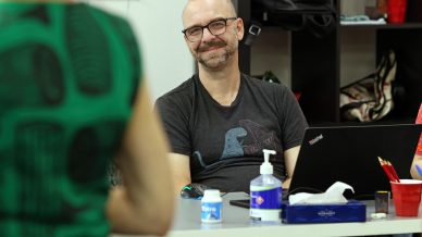 A man with glasses and a moustache smiles while sitting at a desk with a laptop, hand sanitiser, tissues, and a water bottle. Another person stands in the foreground, partially out of focus.
