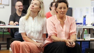 Two women sit close together on a bench, one looking up thoughtfully and the other gazing forward with a serious expression. People and office items are visible in the background, suggesting a casual or collaborative setting.