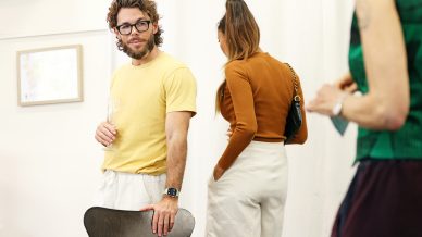 A man in a yellow shirt and glasses holds a champagne glass and stands by a chair, whilst two women nearby talk together. The setting appears to be a casual indoor gathering.