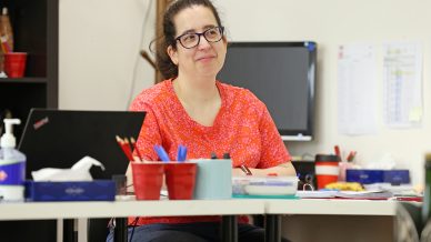 A woman wearing glasses and a red patterned blouse sits at a desk cluttered with office supplies, smiling thoughtfully. Behind her is a computer monitor and various office items.