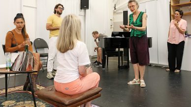 A group of six adults are gathered in a brightly lit room. One person stands and gestures expressively, while others sit or stand around, listening and interacting. There is a piano and a rug in the room.