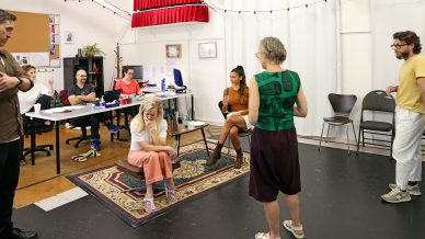 A group of people gather in a bright, casual office space. Some sit and others stand, engaged in conversation. Office desks, chairs, and computers are in the background, and a rug decorates the floor.