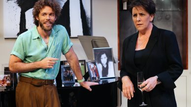 A man in a green shirt and brown trousers stands beside a piano covered with framed photos, smiling. A woman in a black suit stands beside him, holding a glass, looking concerned. Modern art hangs on the wall behind them.