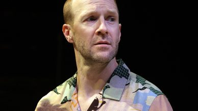 A man with short hair and a trimmed beard wears a colourful, patterned short-sleeve shirt with palm trees and holds a drink, looking thoughtfully to the side against a dark background.