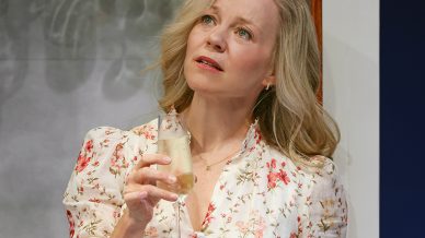 A woman with blonde hair, wearing a white floral dress, stands in front of a drinks cabinet holding a glass of champagne, looking upwards with a thoughtful expression. Bottles and glasses are visible behind her.