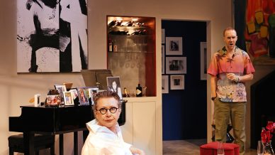 A woman with glasses sits on a white chair in a modern living room, looking at the camera. A man stands in the background. The room has a piano, family photos, abstract art, and a white rug.