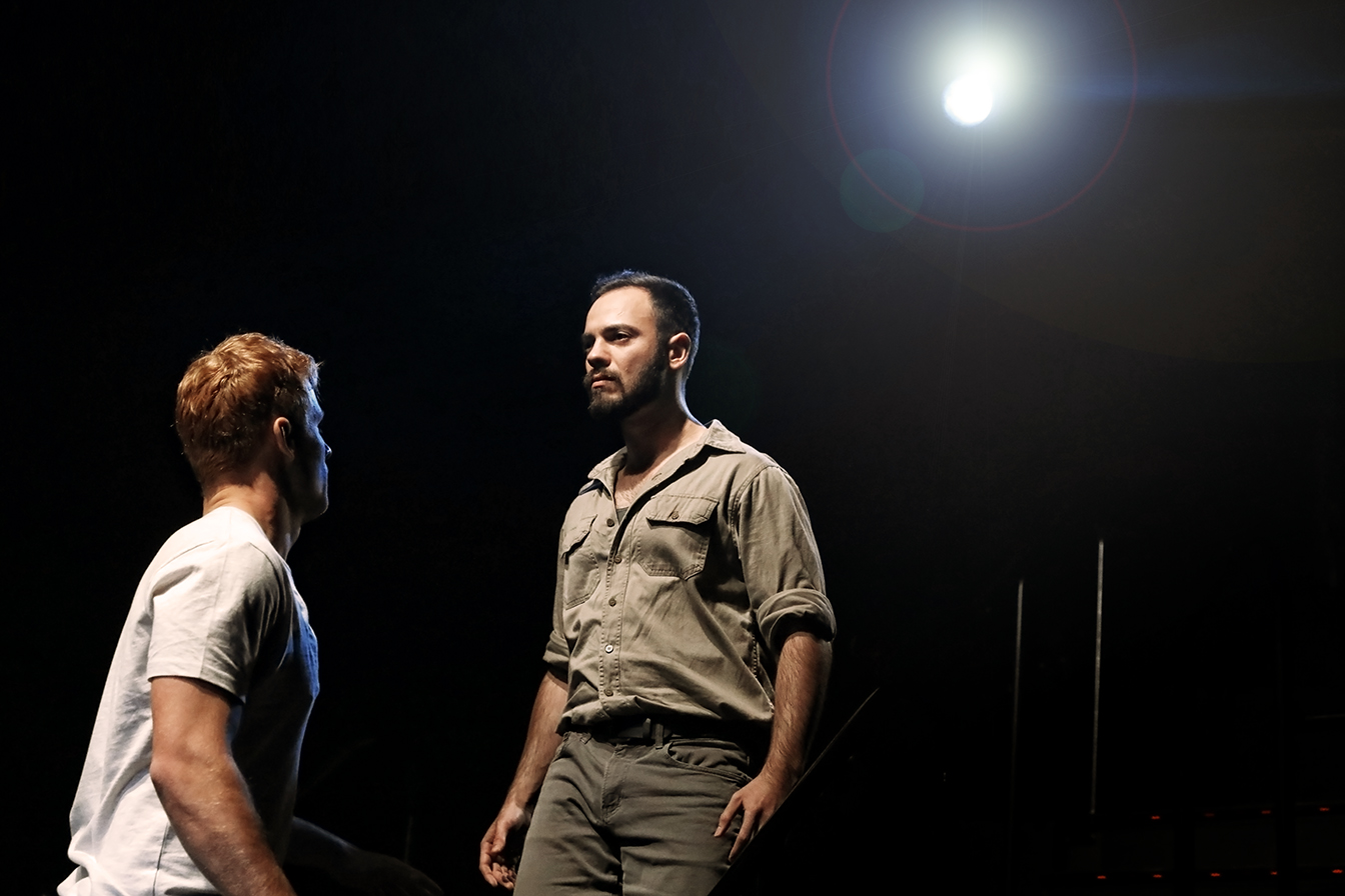 Two men face each other intensely on a dark stage under a bright spotlight, one wearing a white T-shirt and the other in a khaki shirt, creating a dramatic atmosphere.
