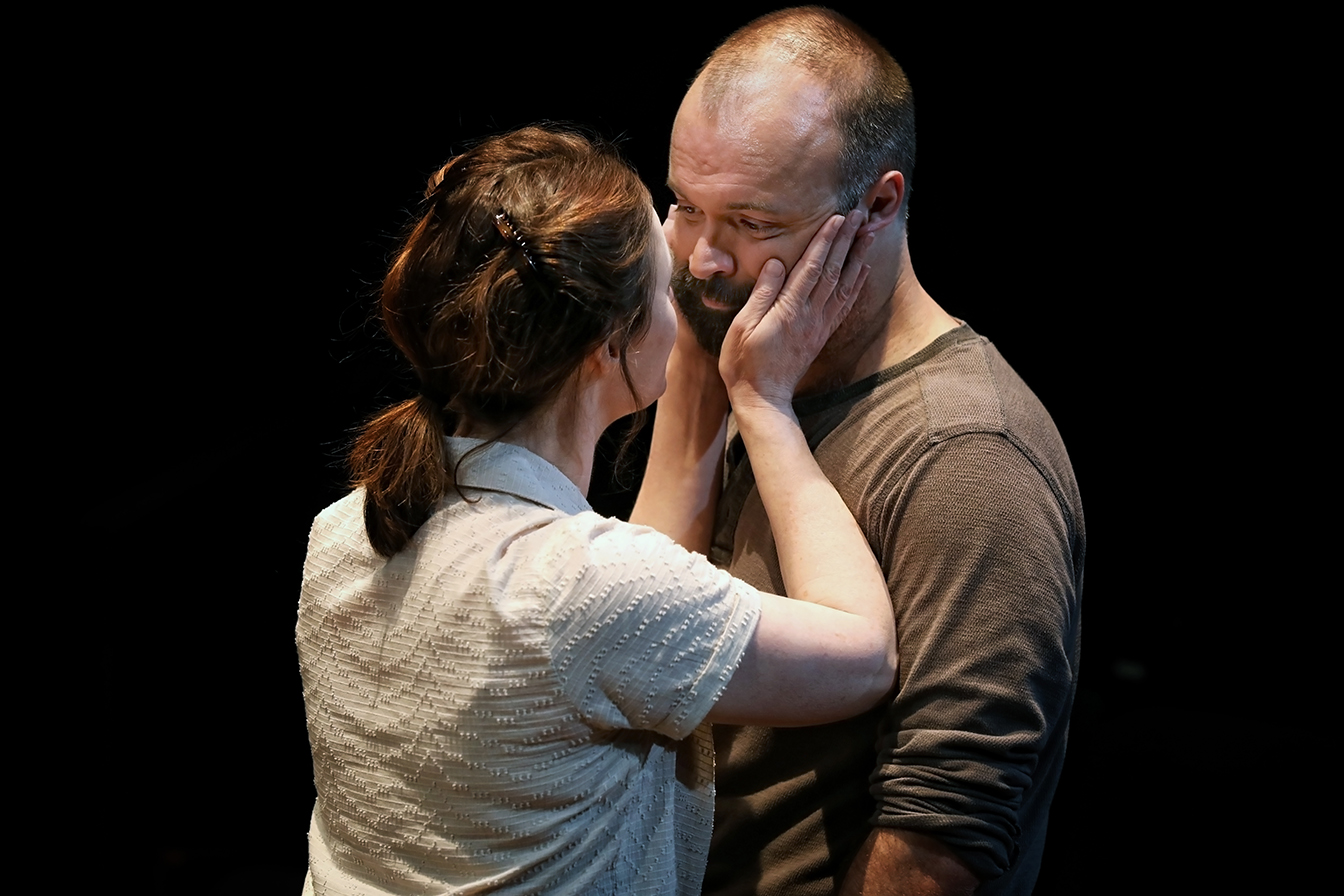 A woman gently holds a man's face in her hands and looks into his eyes. The man has a beard and appears emotional. They are standing close together against a dark background.