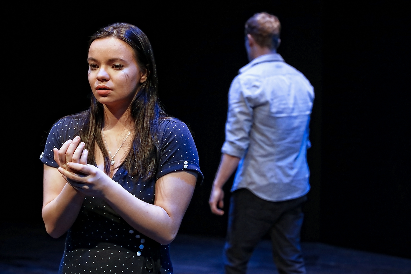 A woman stands in the foreground looking upset and holding her hands together, whilst a man stands in the background with his back turned, both on a dark stage.