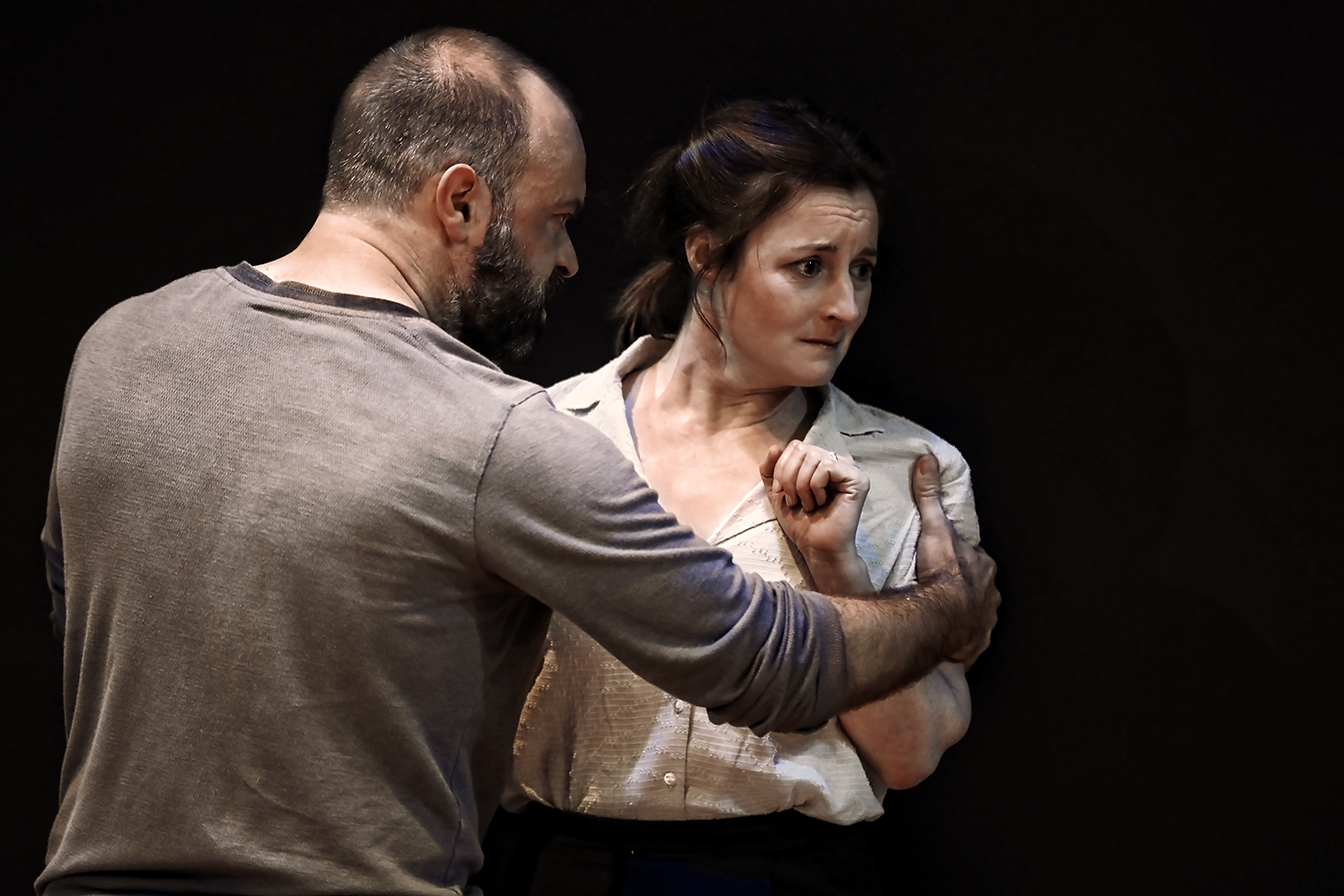 A man holds a woman by the arm, his expression intense, while the woman looks anxious and recoils slightly, clutching her hands to her chest against a dark background.