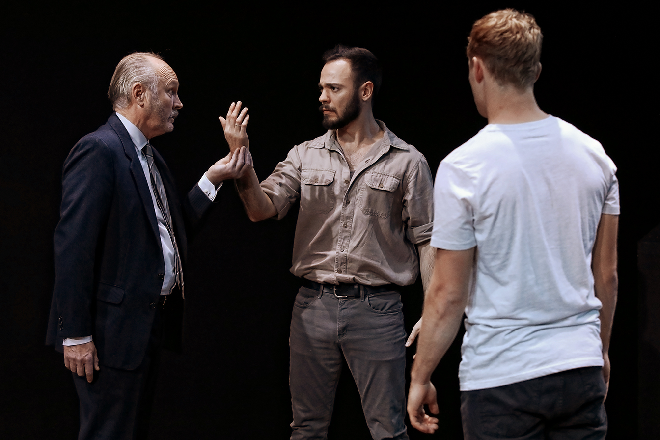 Three men are on a dark stage; one older man in a suit faces a younger man in casual clothes who holds up his hand between them, while a third man in a white T-shirt stands with his back to the camera, watching.