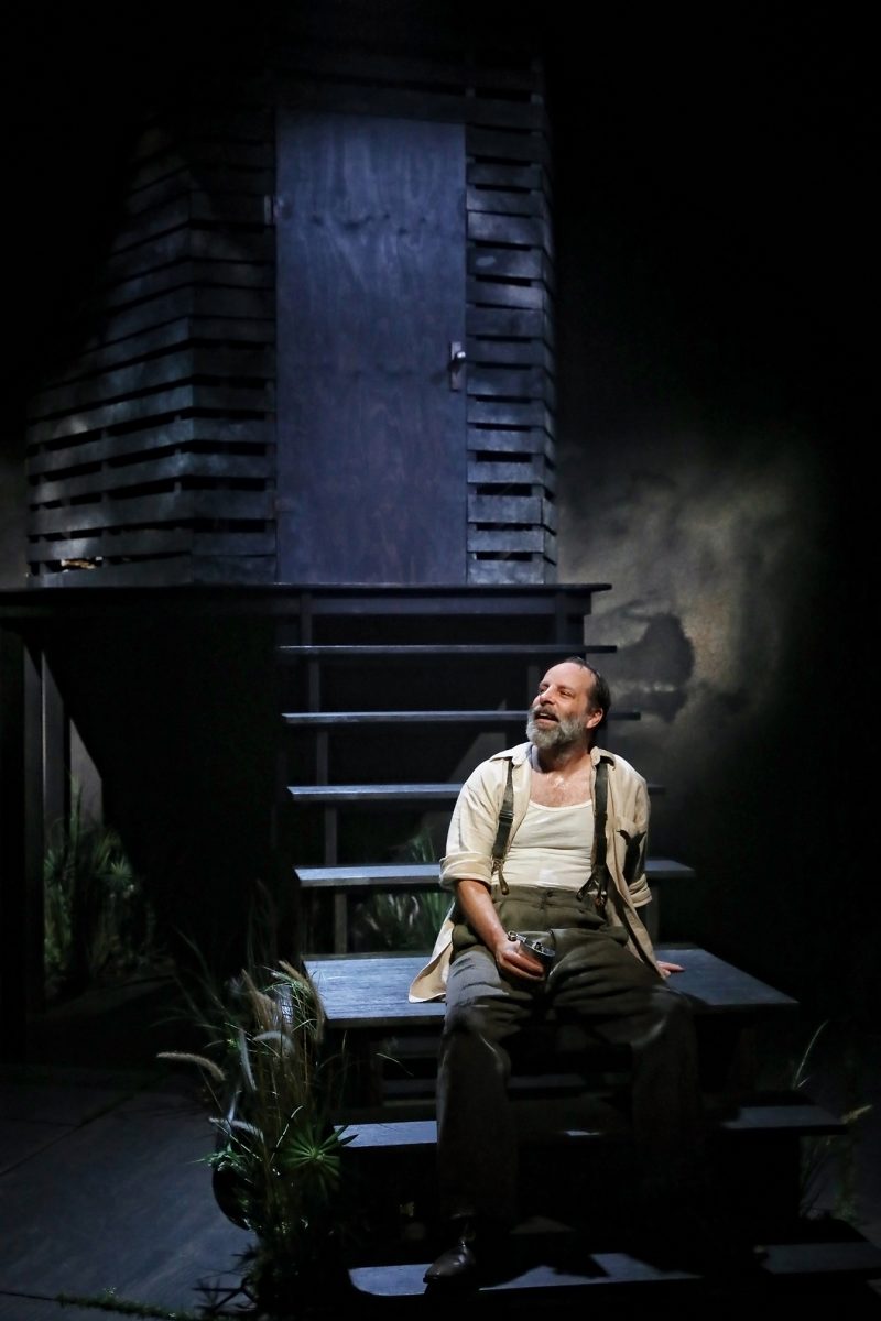 A bearded man in braces and a white shirt sits on outdoor steps, holding a small object, in front of a dark wooden structure under dramatic stage lighting.
