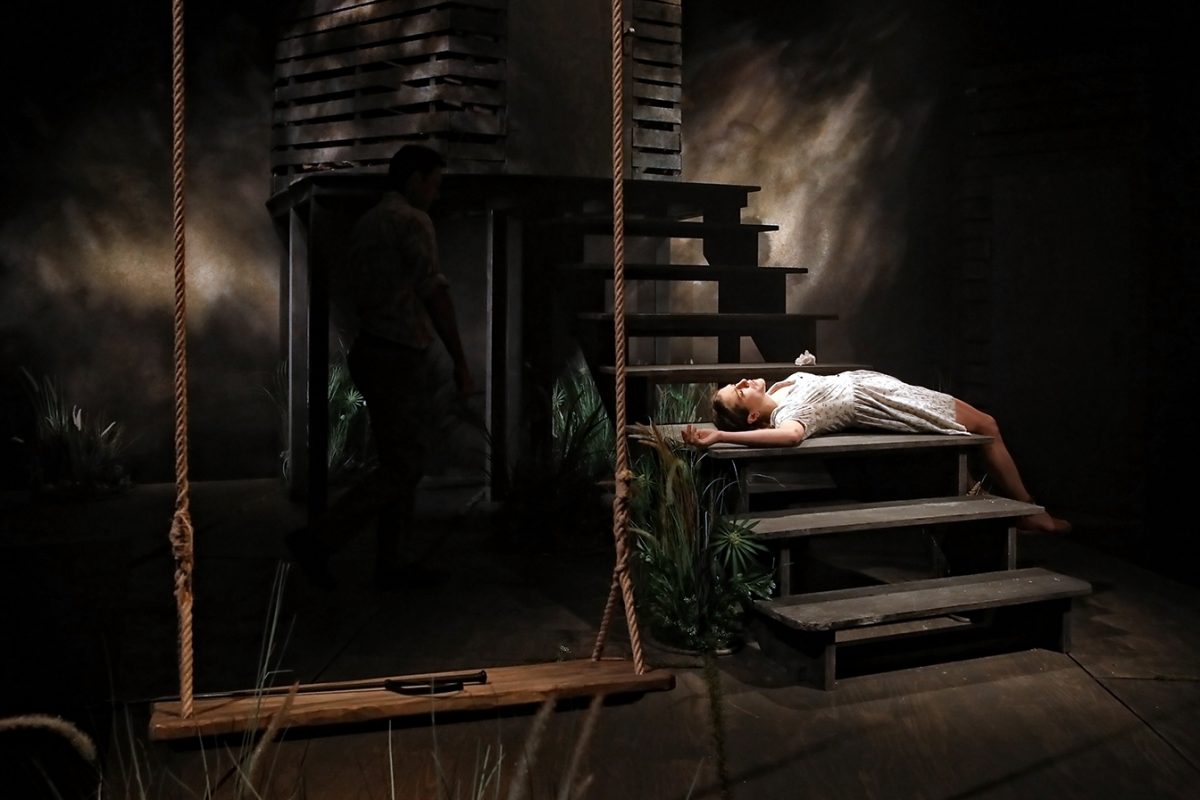 A woman in a white dress lies on her back across wooden stairs, illuminated by a spotlight. A man in shadow stands nearby. A large rope swing hangs in the foreground of the dimly lit, rustic setting.