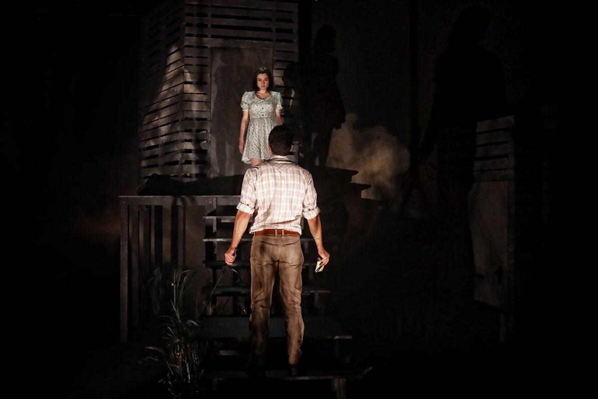 A man stands at the bottom of wooden stairs, facing a woman in a light dress who stands at the top. The scene is dimly lit, with dramatic shadows and mist creating a tense, mysterious atmosphere.