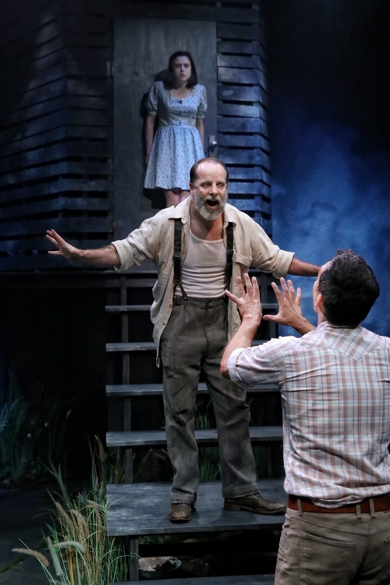 A bearded man in braces stands with arms outstretched on wooden steps, facing another man with raised hands. A worried woman in a dress stands in a doorway behind them, watching the scene. The setting appears dramatic and tense.