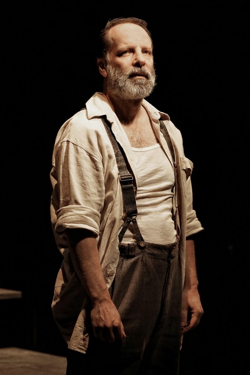 A bearded man stands under dramatic lighting, wearing a white shirt with rolled-up sleeves, braces, and dark trousers, looking slightly upward with a serious expression. The background is dark and shadowy.