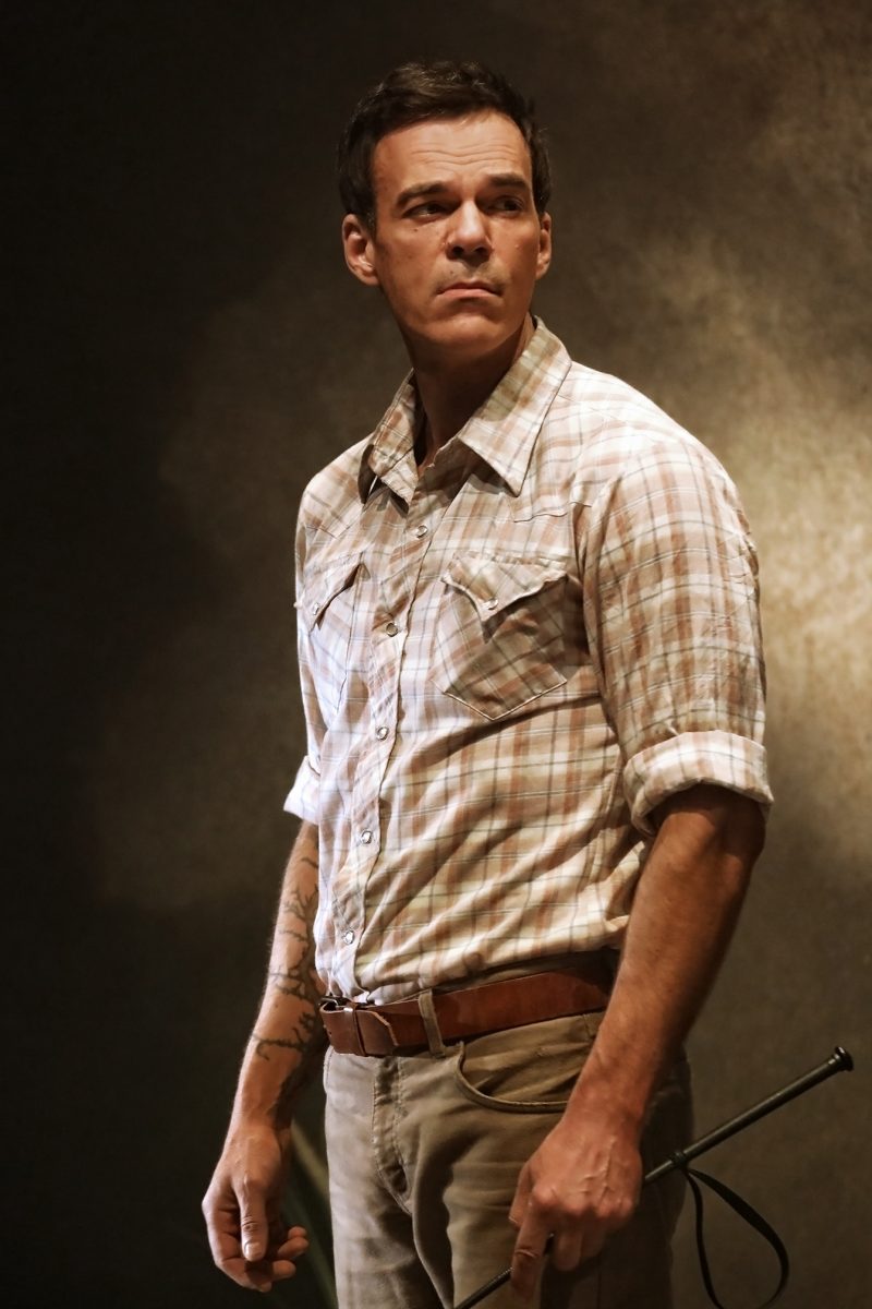 A man wearing a checked shirt and brown belt stands against a dark, textured background, holding a riding whip. He gazes to the side with a serious expression.