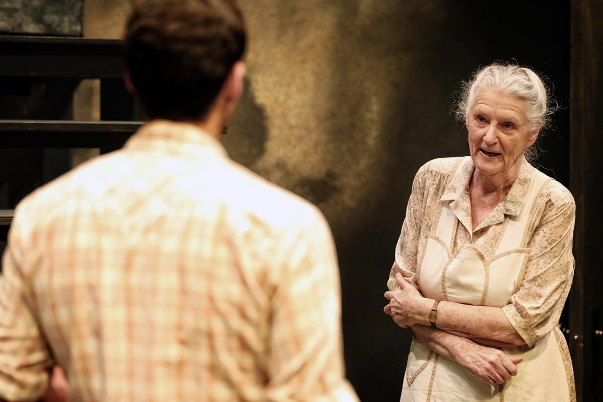 An older woman with grey hair in a bun and wearing a beige dress stands with her arms folded, speaking to a younger man whose back is to the camera. The scene appears to be on a theatre stage.