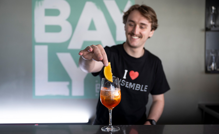 A smiling barman garnishes a cocktail with an orange slice, standing behind a bar with a large BAYLY sign in the background.