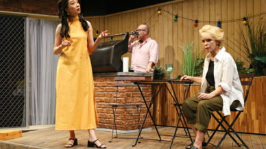 Three people are on a wooden decking set; a woman in a yellow dress stands speaking, a seated woman in casual clothes listens, and a man in sunglasses drinks behind them. The setting is decorated with fairy lights and plants.