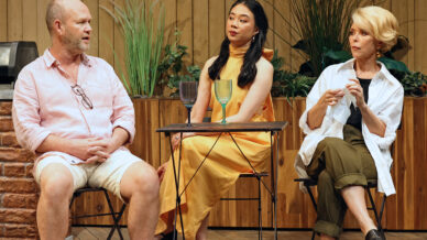 Three adults sit on garden chairs around a small table, engaged in conversation. The man on the left wears a light pink shirt and shorts, while the two women wear a yellow dress and white shirt with green trousers. Plants decorate the background.