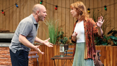 A man and a woman are arguing intensely beside a table with a wine bottle and glasses, in an outdoor setting with fairy lights, plants, and wooden fencing. The woman holds up her hand defensively.