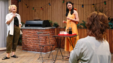 Three women are on a wooden patio set. One woman in a white shirt laughs on the left, another in a yellow dress looks confused at centre, and a third with her back to the camera faces them. Fairy lights hang in the background.