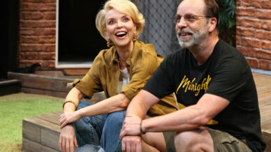 Two people, a smiling woman in a brown shirt and jeans and a man in glasses and a black Midnight shirt, sit side by side on wooden steps outdoors, appearing relaxed and happy.