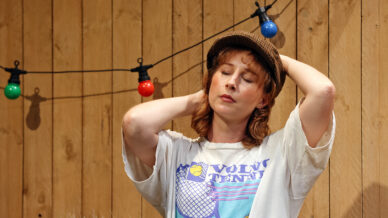 A person wearing a brown cap and a VOLVO TENNIS T-shirt stands in front of a wooden fence, eyes closed, hands behind their head, with colourful fairy lights hanging in the background.