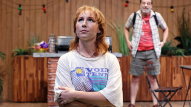 A woman with short red hair and a VOLTENNIS T-shirt stands with arms folded, looking up thoughtfully. Behind her, a man in casual clothes with a rucksack stands near outdoor patio furniture and wooden decor.