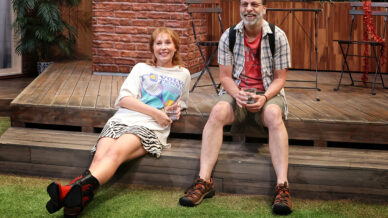 A woman and a man sit on wooden steps outside, smiling and holding drinks. The woman wears a white T-shirt and striped skirt, while the man wears a checked shirt and shorts. They are surrounded by plants and a wooden decking.
