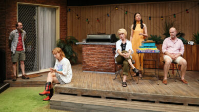 Five people are on a wooden patio set. Two sit on the decking, one stands by a door, one sits near stacked board games, and one stands speaking. Colourful fairy lights hang on the fence in the background.