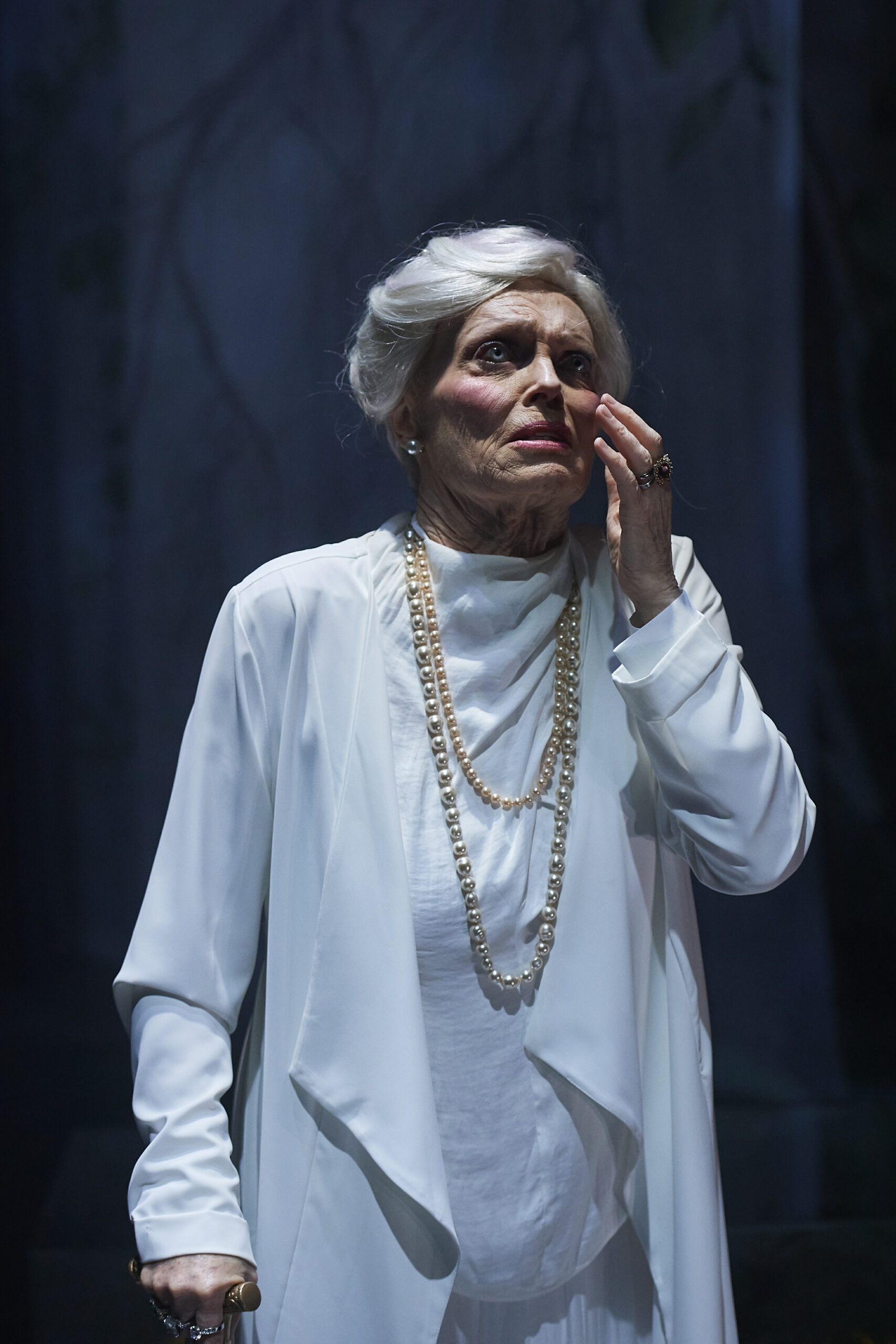 An elderly woman with white hair, wearing a long white outfit and pearl necklaces, stands with a concerned expression, touching her face with one hand and holding a walking stick with the other, in a dimly lit setting.