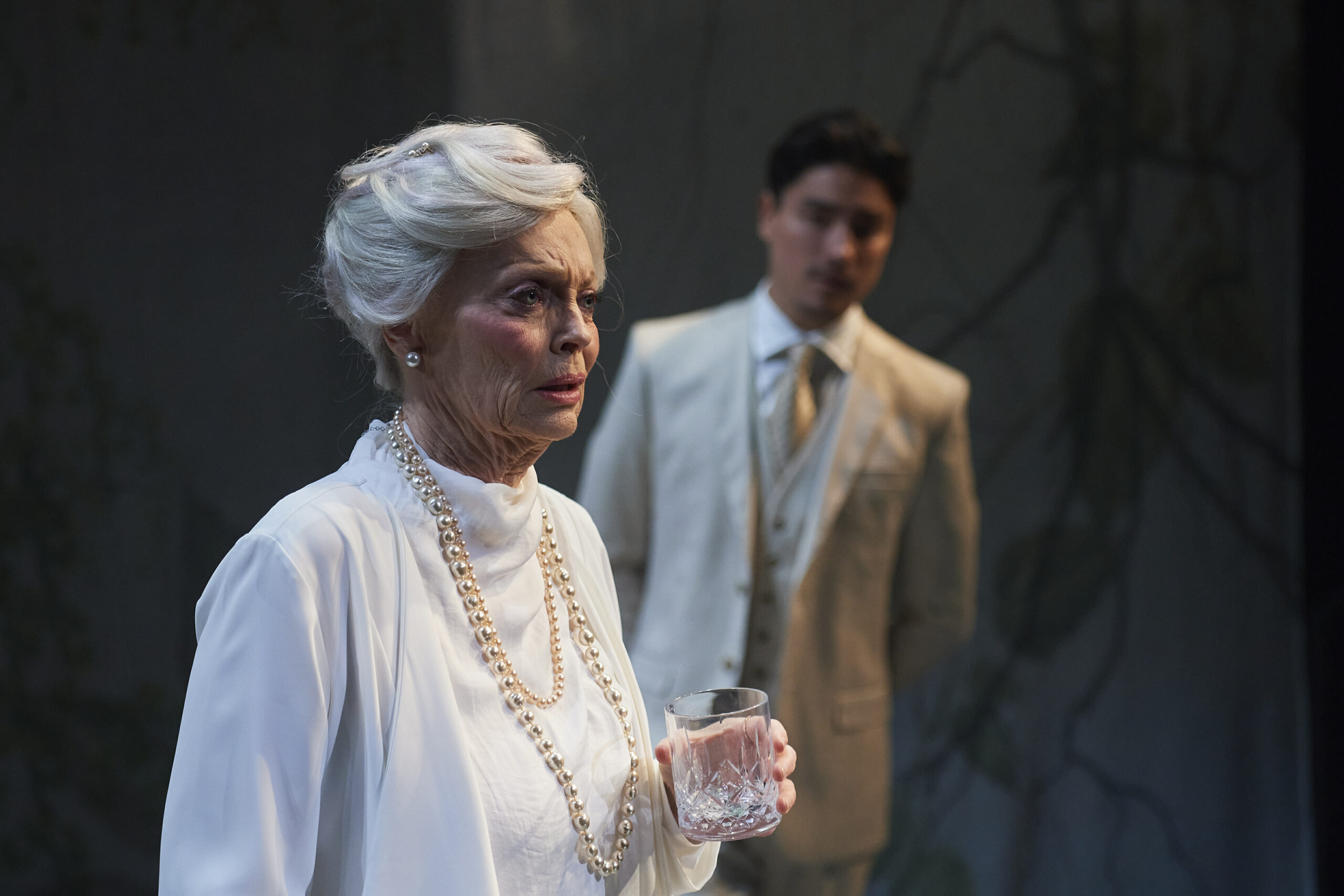 An elderly woman with white hair and pearl necklace holds a glass and looks serious, whilst a younger man in a light suit stands blurred in the background. The scene appears dramatic and tense.