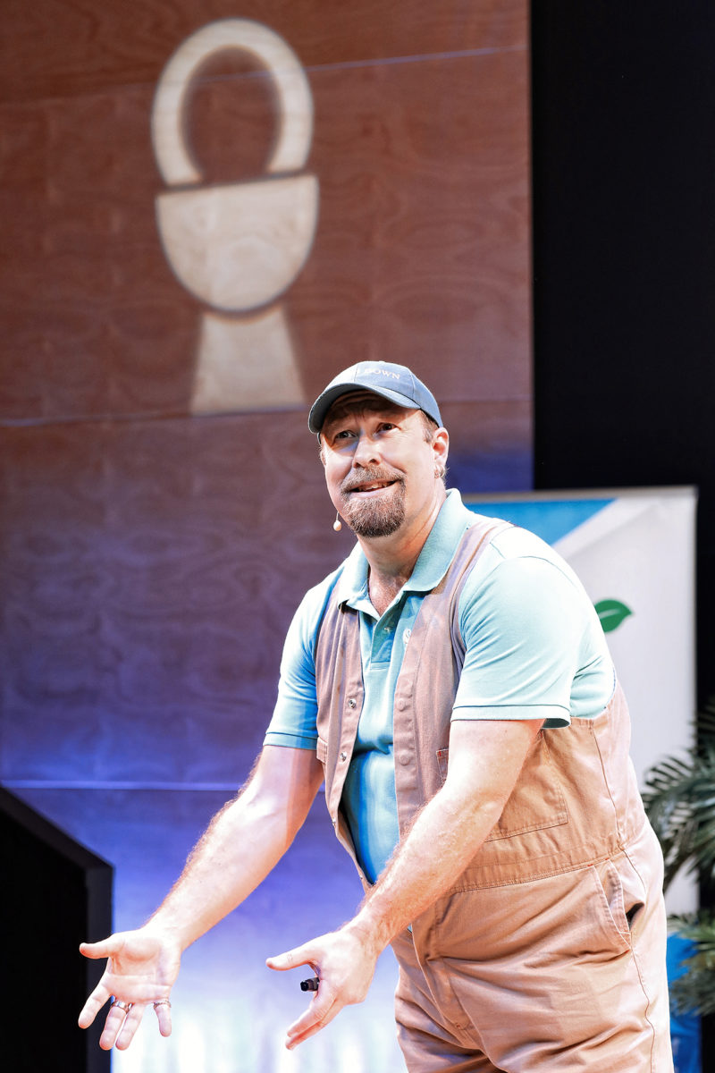 A man in a brown waistcoat and blue shirt stands onstage, gesturing with his hands. He wears a cap and has a goatee. Behind him, a spotlight projects a lock symbol onto a wooden wall.