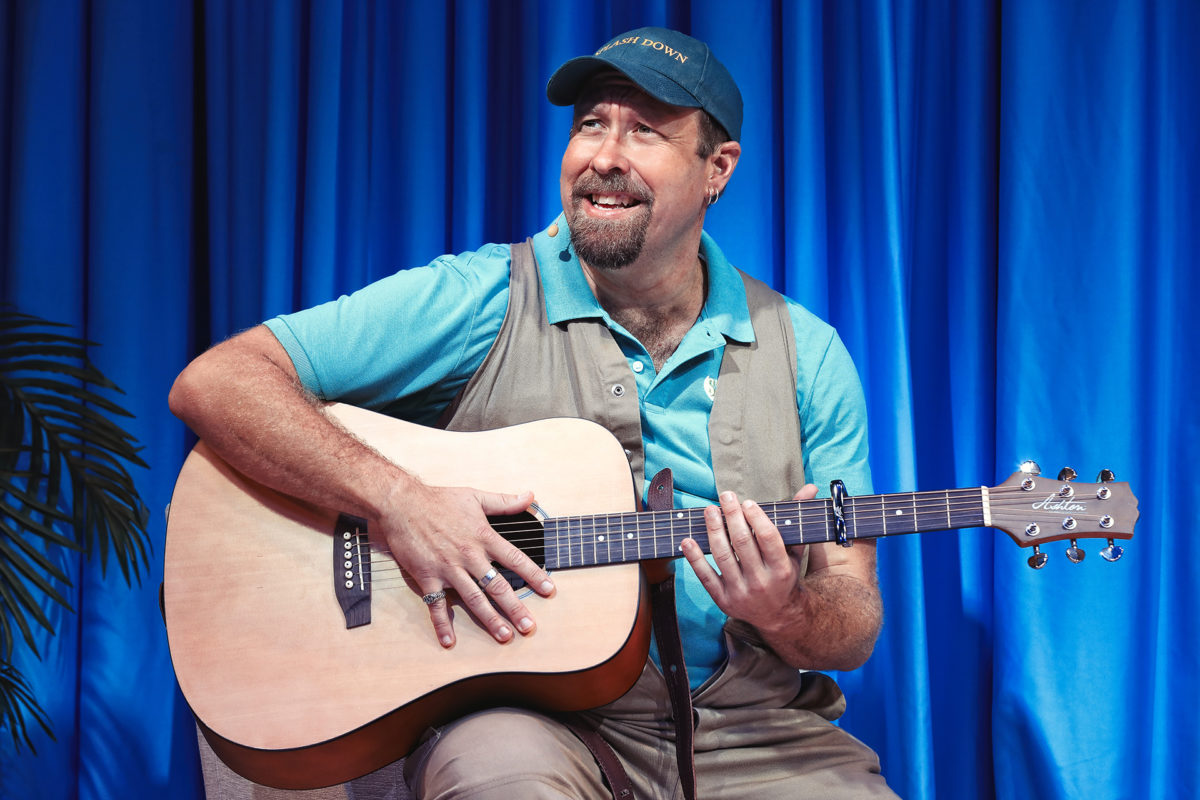 A smiling man in a blue cap and shirt plays an acoustic guitar on stage, seated in front of blue curtains, with a plant partially visible on the left.