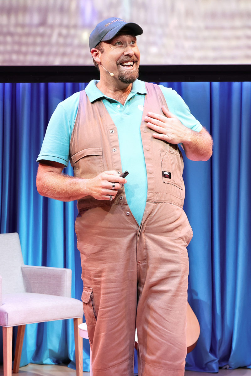 A man wearing a cap, blue polo shirt, and tan dungarees stands on stage smiling with one hand on his chest, holding a small object in the other hand. A white chair and a blue curtain are in the background.