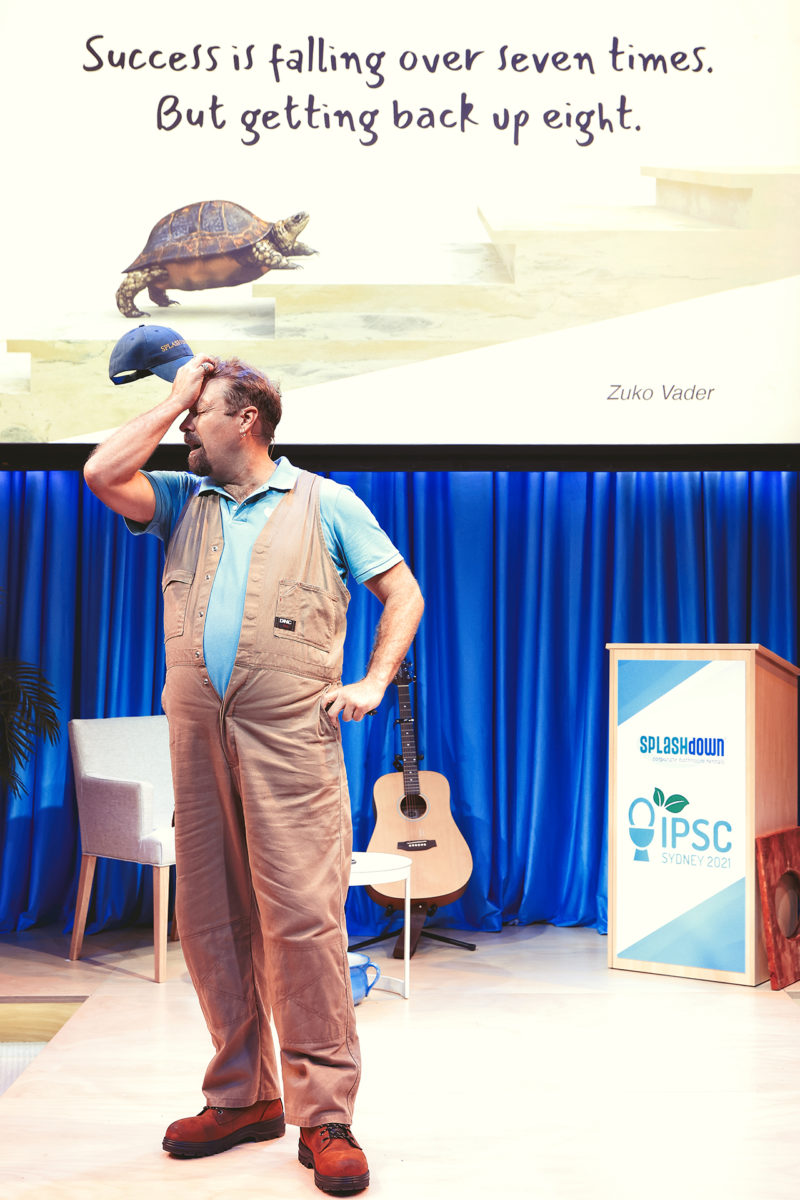 A man in work overalls stands on stage, tipping his cap, with a guitar and a lectern nearby. Behind him, a slide shows a tortoise next to the quote: Success is falling over seven times. But getting back up eight.