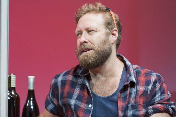 A bearded man in a checked shirt and blue vest sits in front of a reddish-pink background, with two wine bottles beside him. He looks off to the side with a thoughtful expression.